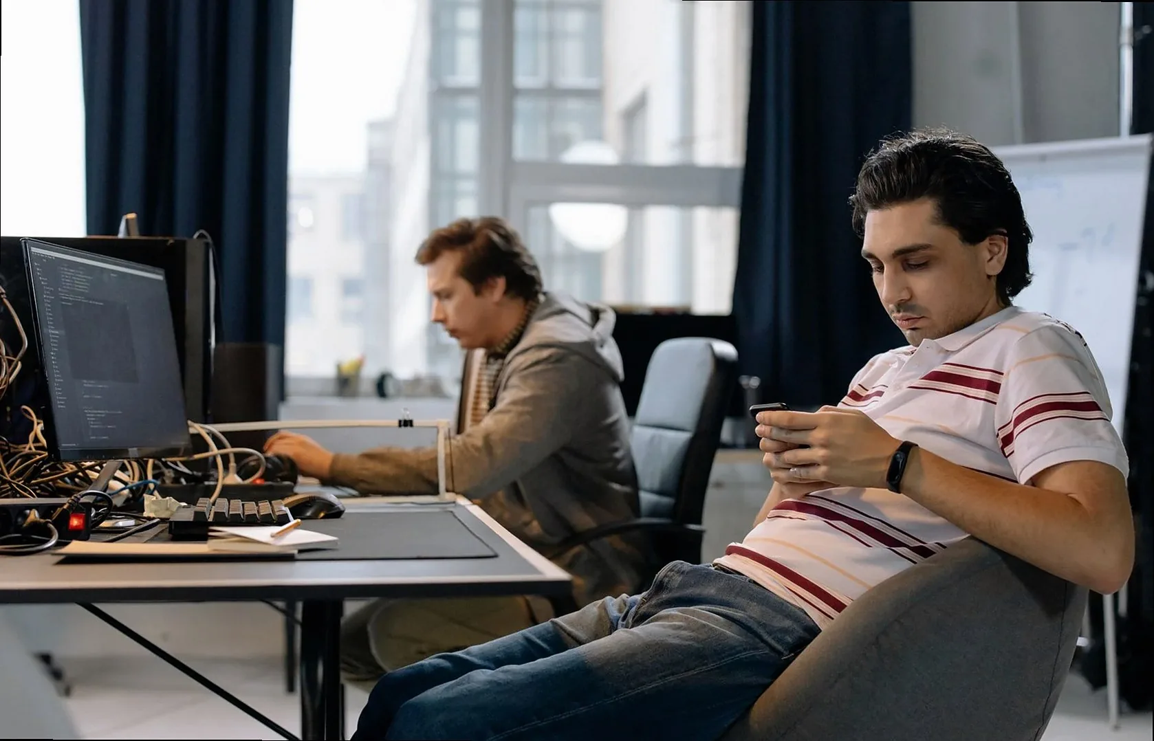 Two men working in a modern office, one on a computer and the other on a smartphone.
