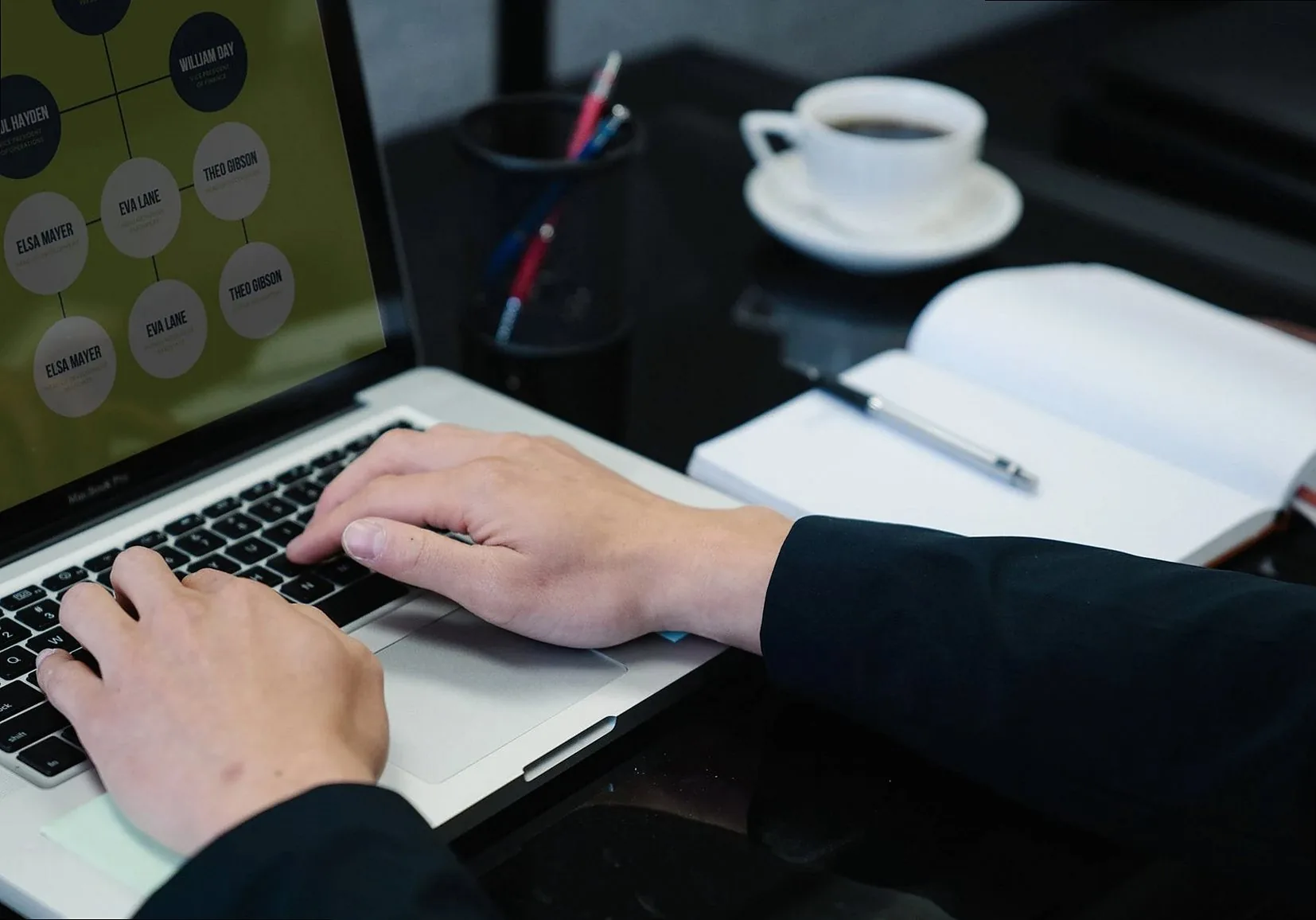 Close-up of hands typing on a laptop displaying an organizational chart, a notebook, and coffee nearby.